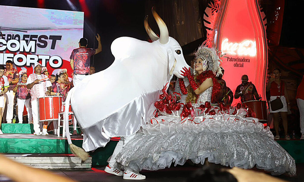 Fotografia horizontal e festiva do palco iluminado no Sambódromo de Manaus durante a Noite da Celebração Folclórica. Ao centro, as cantoras Gaby Amarantos e Márcia Siqueira dividem o microfone sorridentes, cercadas pelos itens oficiais do Boi Garantido e dançarinos do Garantido Show com trajes folclóricos vermelhos e brancos. Ao fundo, a Batucada toca seus tambores sob uma chuva de confetes vermelhos e efeitos de luz, enquanto uma multidão entusiasmada agita bandeiras e braços no compasso da toada.