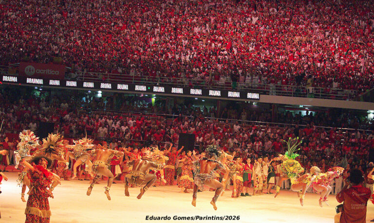 Fotografia horizontal em plano médio na arena do Bumbódromo de Parintins durante o festival de 2026. No centro do palco, integrantes do Garantido Show e do Balé Folclórico da Bahia realizam uma coreografia expressiva e enérgica, com os braços erguidos e figurinos que exaltam as raízes africanas. Ao fundo, ergue-se uma imponente alegoria vermelha e branca com formas místicas que misturam elementos indígenas e a figura de uma onça. Detalhes de efeitos de iluminação cênica incidem sobre os dançarinos sob uma leve chuva que brilha no ar. Na arquibancada ao fundo, a galera vermelha agita adereços em uma massa compacta e festiva.